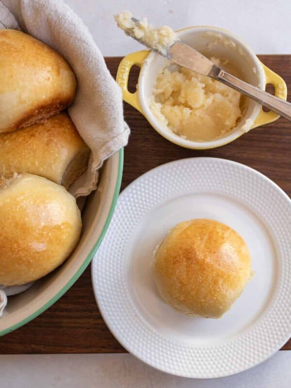 golden sourdough roll on white plate with small yellow dish with butter and a knife and bowl of rolls in the background.