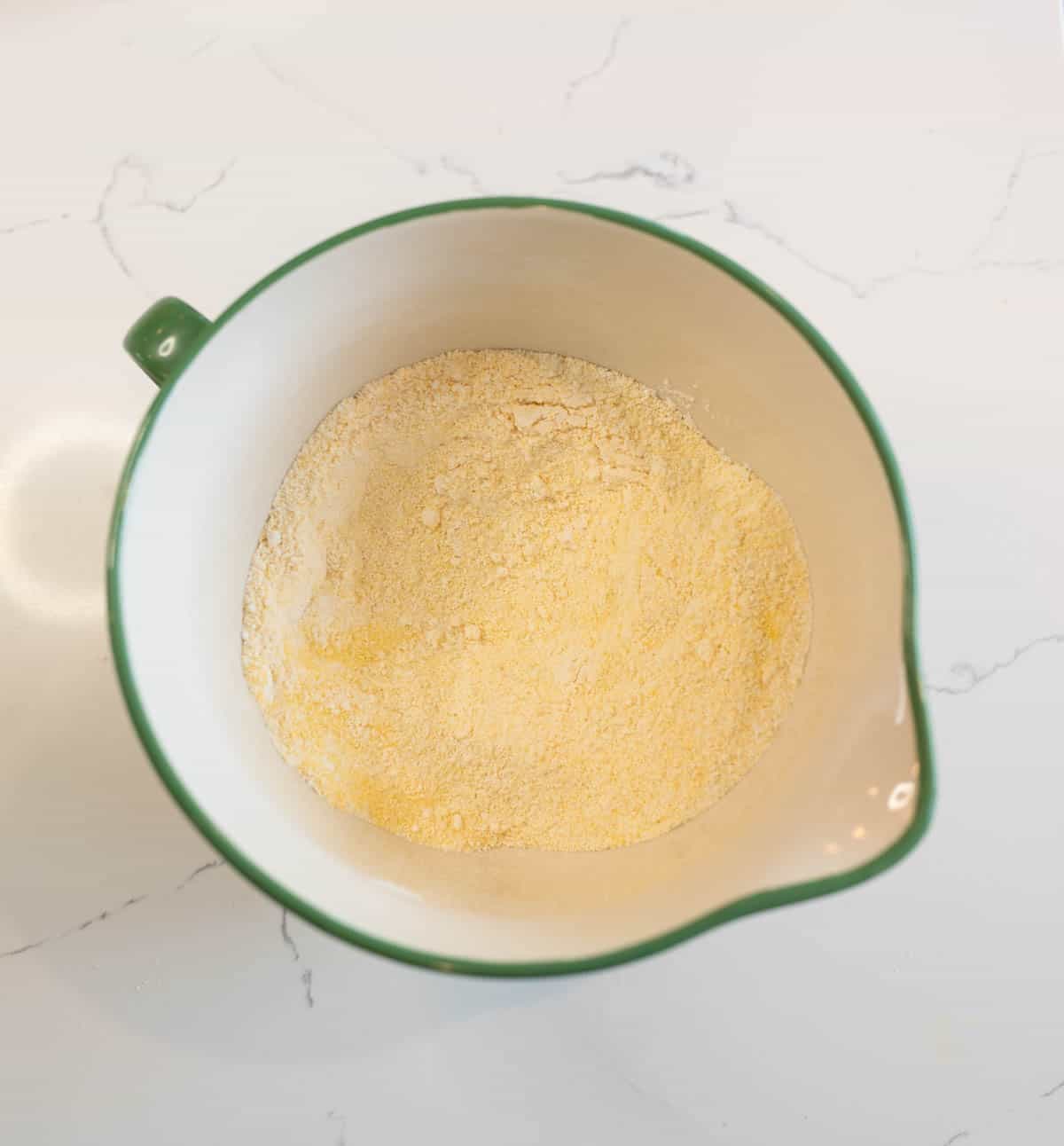 A green-rimmed bowl containing a light yellow powdery substance, likely cornmeal, set on a white marble countertop. The bowls spout is visible at the right side.