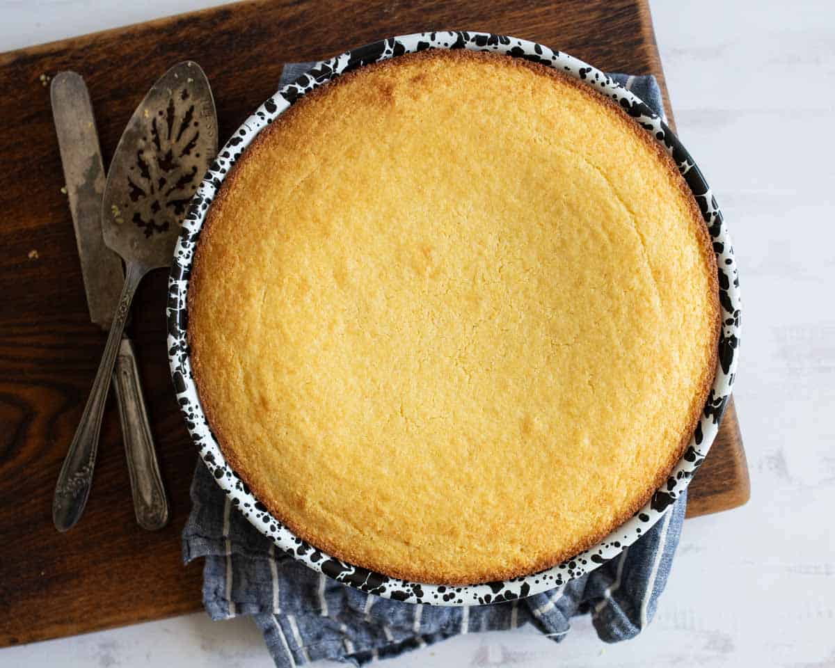 A freshly baked, golden cornbread in a black and white speckled round pan, resting on a wooden board with a gray cloth underneath. Two vintage serving utensils are placed beside the pan. The background is a light-colored surface.