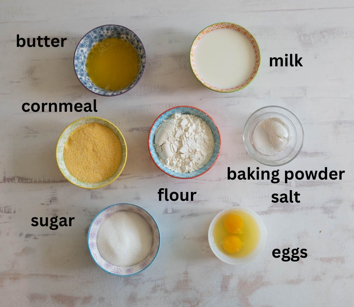 Assorted ingredients in bowls on a white surface: melted butter, milk, cornmeal, flour, baking powder, salt, sugar, and two eggs. Labels are beside each ingredient.