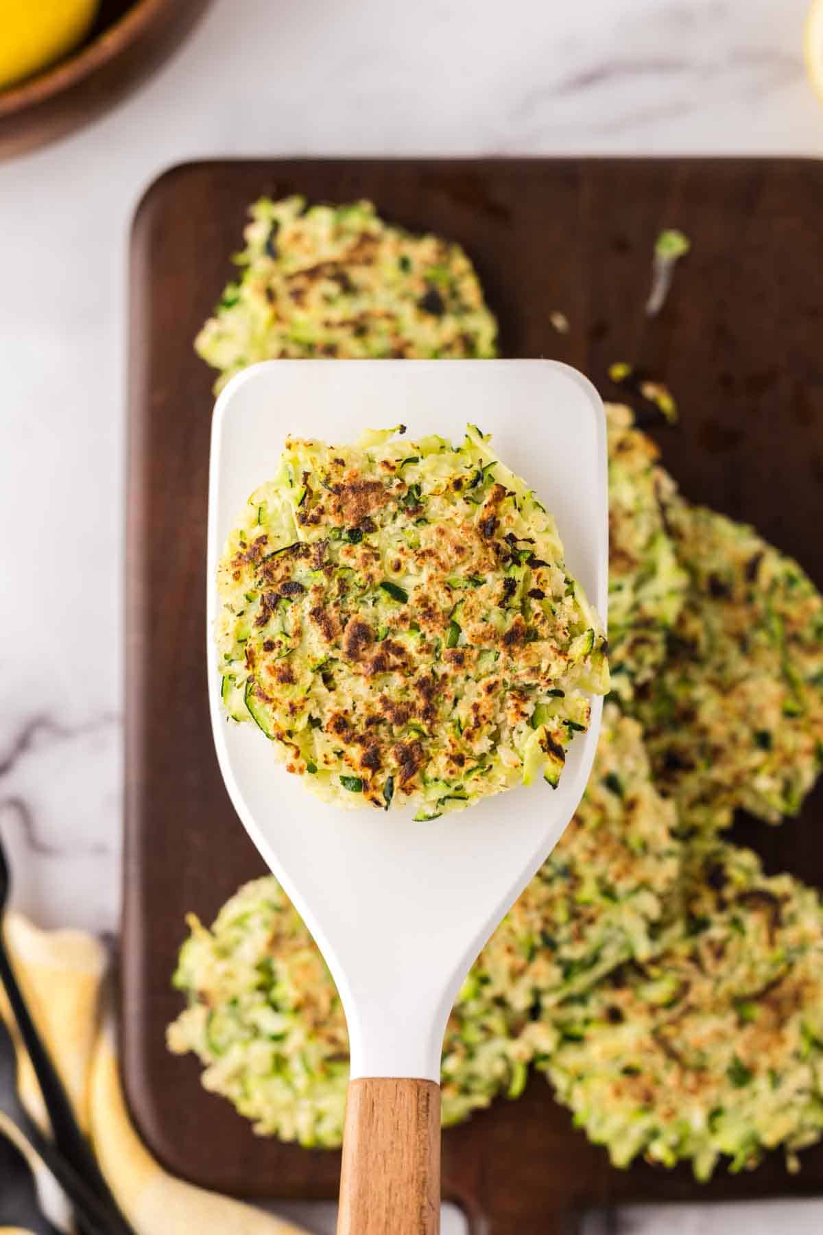 wooden spatula with one fritter over a wooden board with a pile of zucchini fritters.