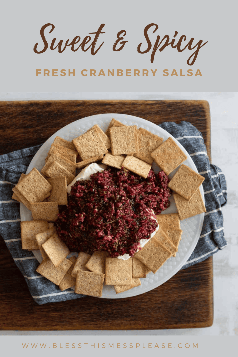 A plate of fresh cranberry salsa sits over cream cheese, surrounded by square crackers. Text above reads Sweet & Spicy Fresh Cranberry Salsa. The image is on a wooden tray with a blue cloth. Website: www.blessthismessplease.com.